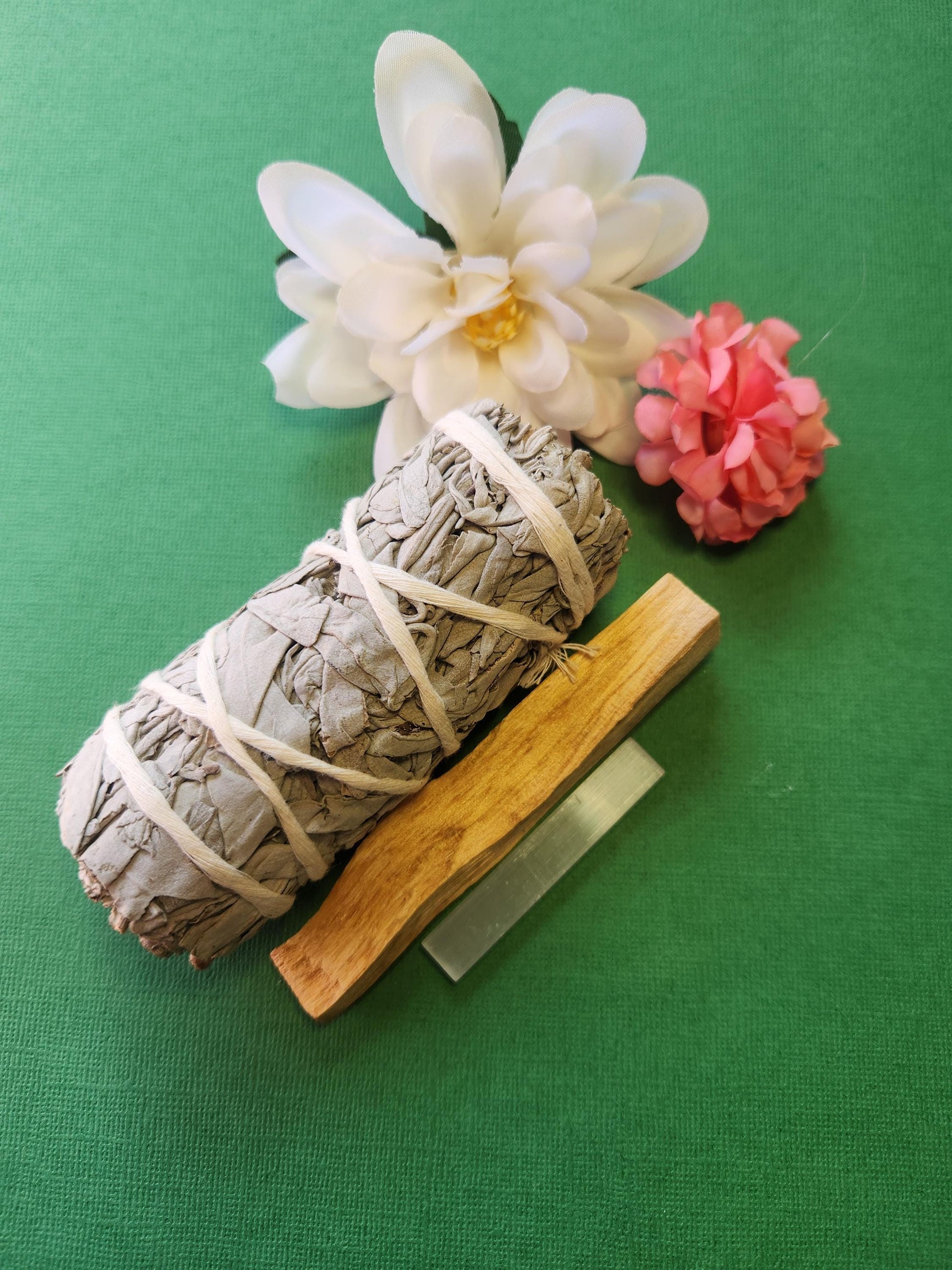 White Sage bundle, Palo Santo stick, and Selenite wand displayed together on a green backdrop with decorative flowers, showcasing a natural cleansing and energy healing set.
