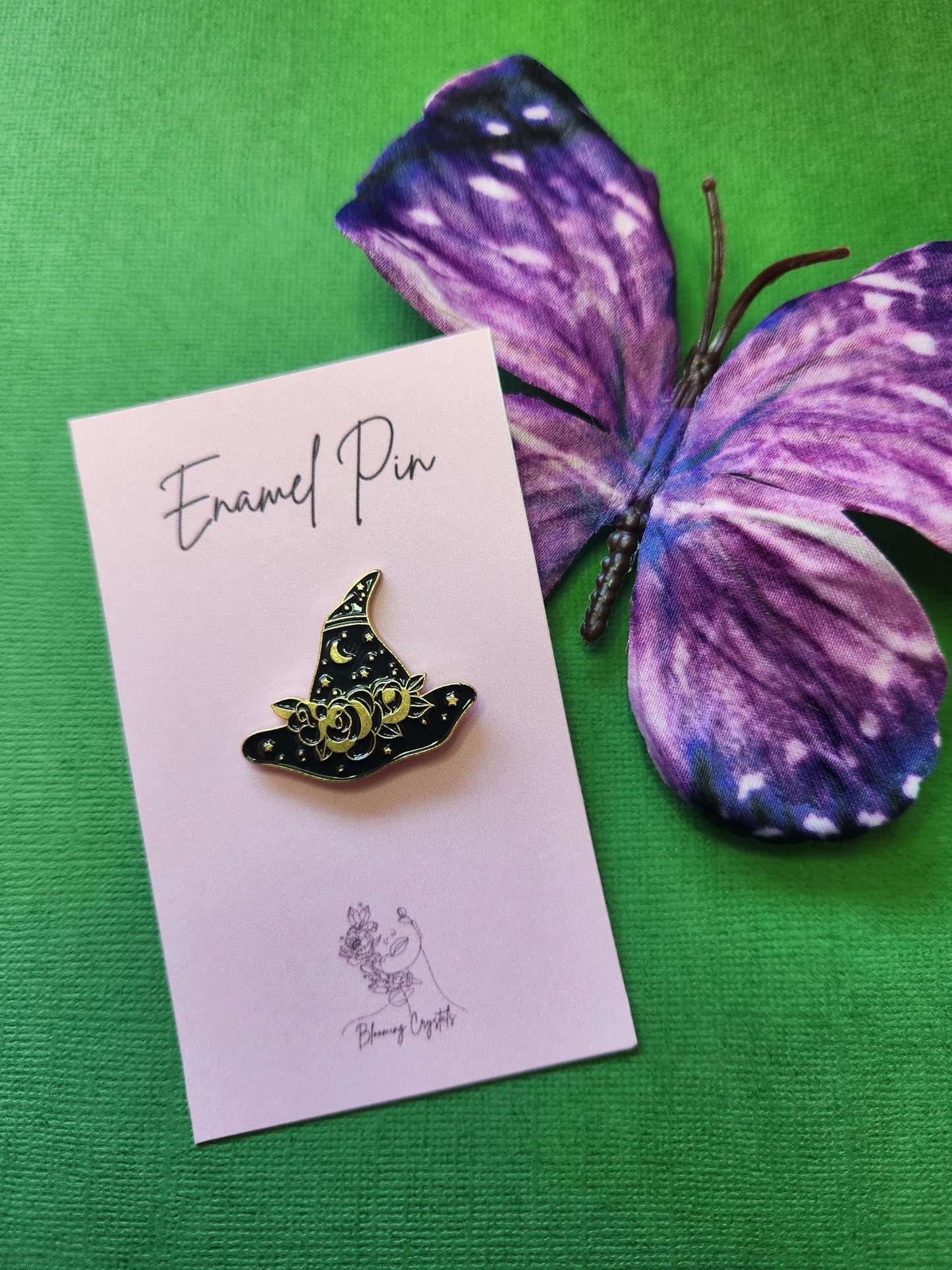 Elegant black enamel witch hat pin adorned with gold floral details, stars, and a small crescent moon. Styled with a purple butterfly prop on a green background. Witch hat enamel pin, celestial witchy accessory, gold and black magical pin, gothic witch fashion, spooky cute Halloween pin.