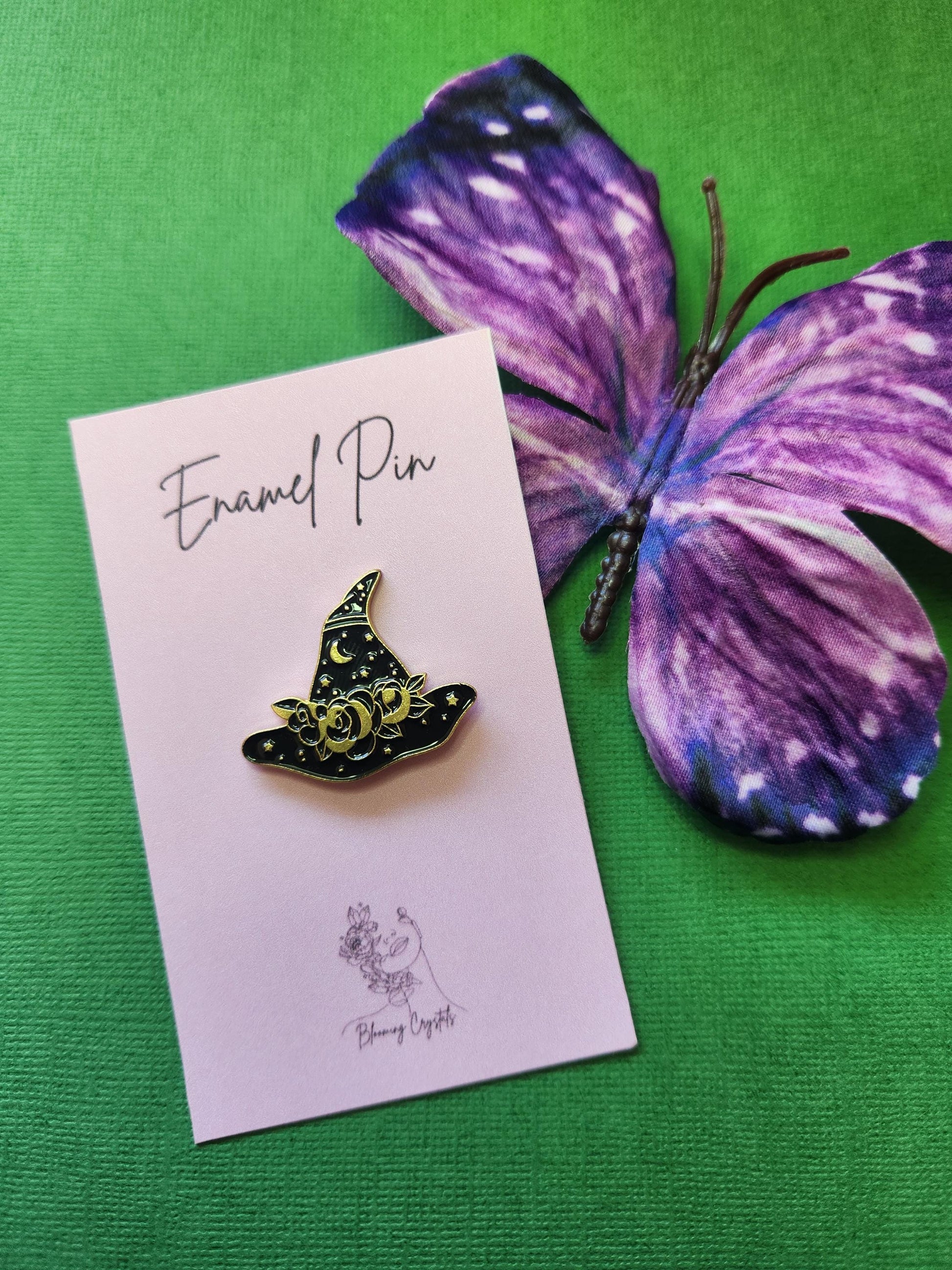 Elegant black enamel witch hat pin adorned with gold floral details, stars, and a small crescent moon. Styled with a purple butterfly prop on a green background. Witch hat enamel pin, celestial witchy accessory, gold and black magical pin, gothic witch fashion, spooky cute Halloween pin.