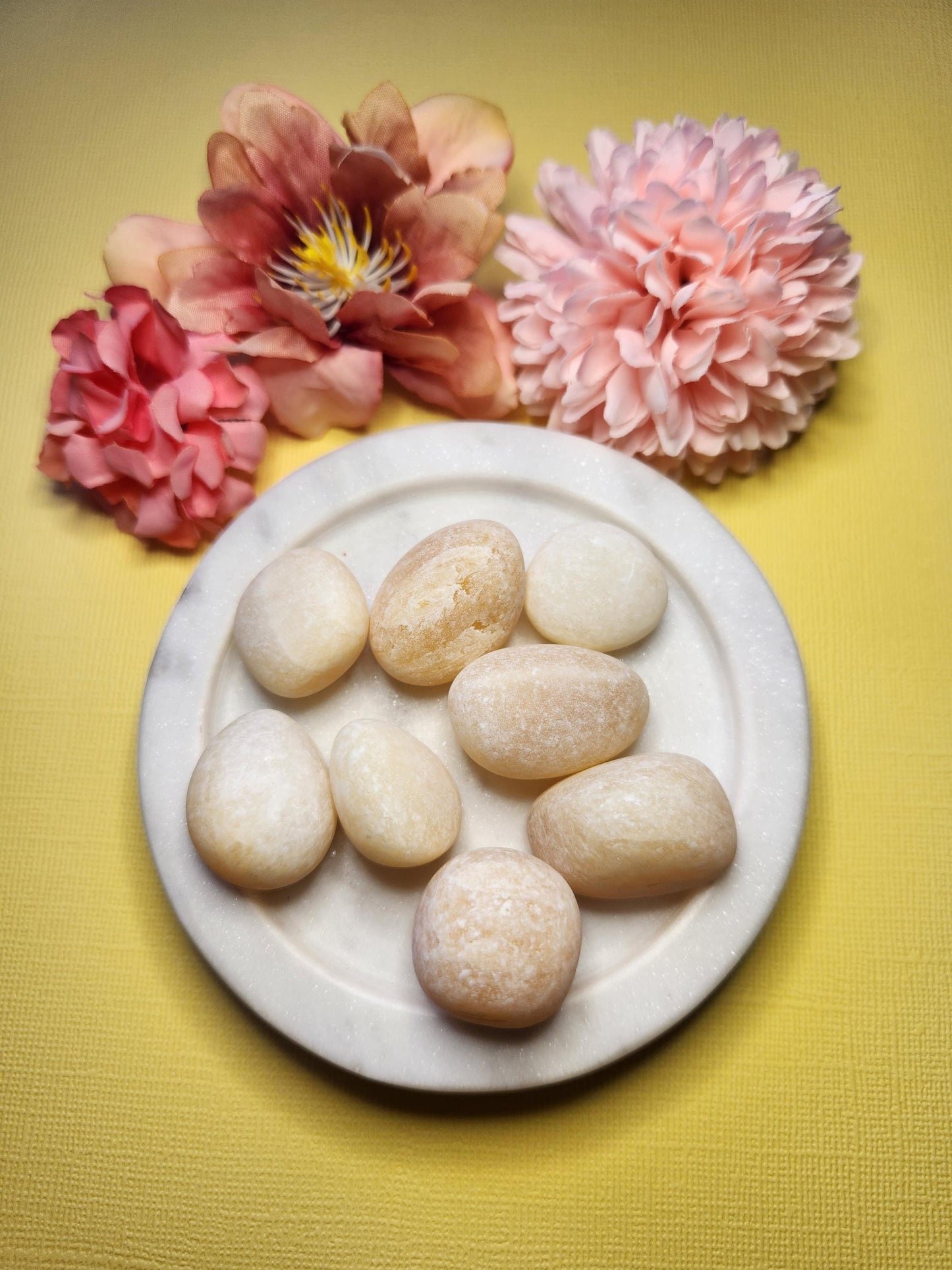 Yellow calcite crystal tumbles on white plate with flowers on yellow background – natural healing stones for energy, joy, and clarity, available in Australia