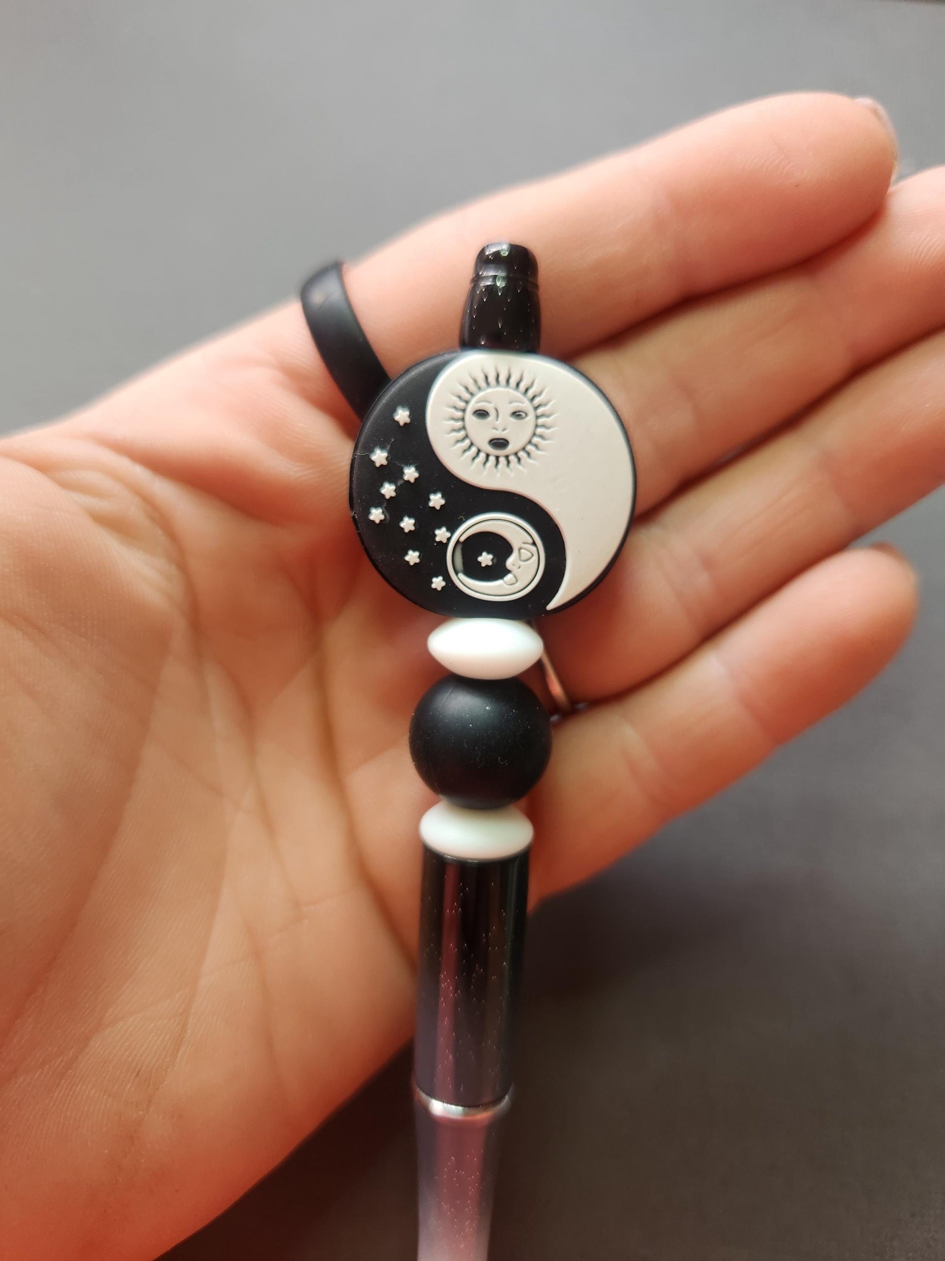 Close-up of Sun and Moon Yin Yang silicone beaded pen held in hand, showing the black and white yin yang focal bead with sun and moon detailing and matching silicone beads on a glitter ombre barrel. High-detail celestial pen, yin yang aesthetic stationery, spiritual writing tool, perfect for journaling, note-taking and gifting.