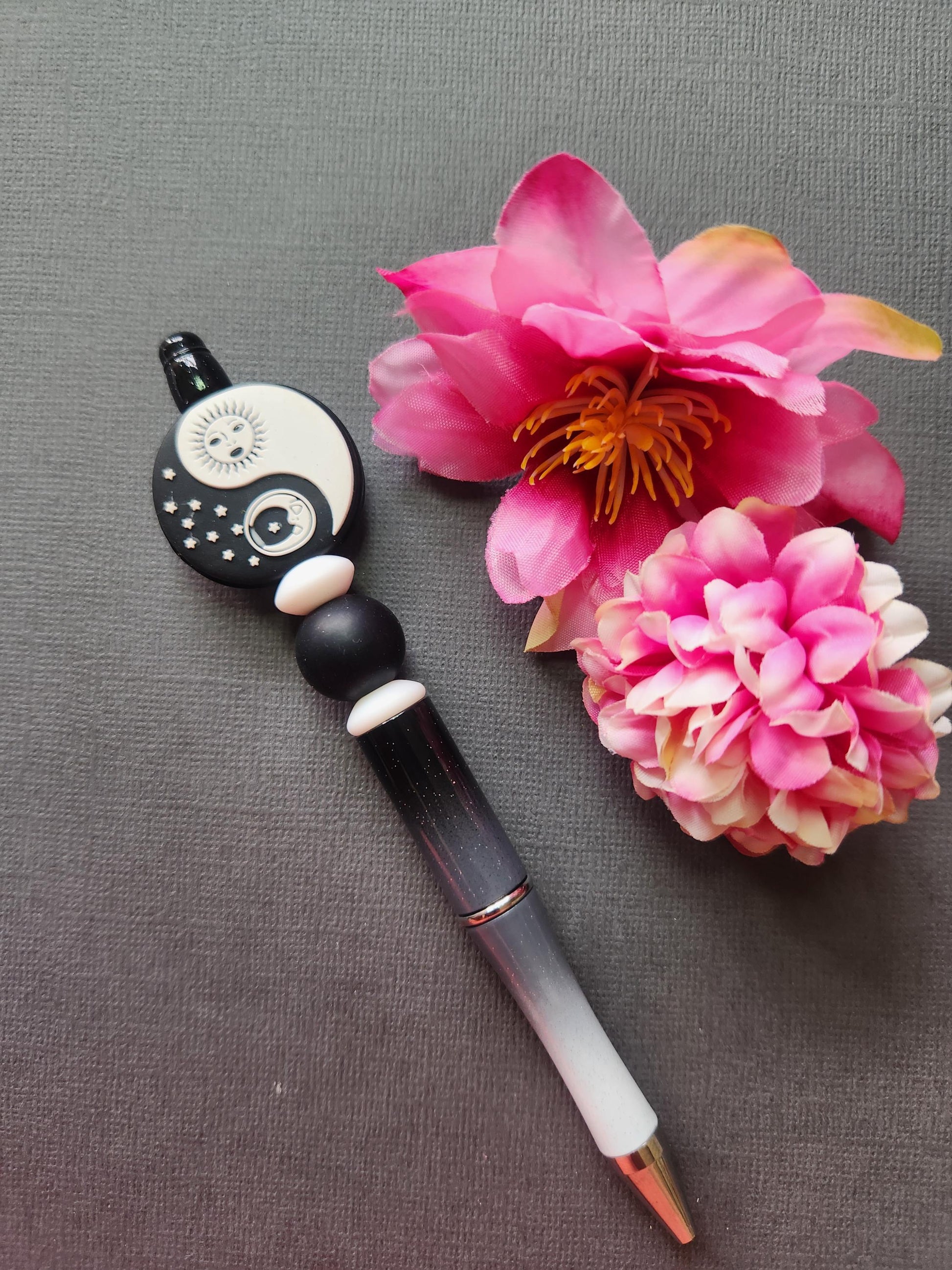 Sun and Moon Yin Yang silicone beaded pen with black and white focal bead featuring celestial sun and moon design, paired with white and black silicone beads on a black-to-white glitter ombre pen barrel. Pen displayed on a dark grey background with two flower props. Aesthetic celestial stationery, yin yang pen, spiritual writer gift, silicone beaded pen for journaling and study